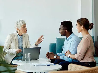 A professional meeting between a consultant and two clients, discussing plans with a laptop and paperwork on a table.