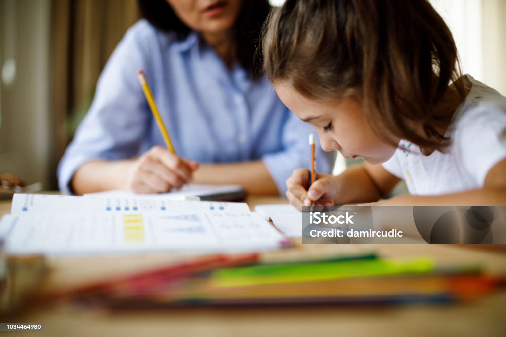 Mother helping daughter with homework A student is engaged in writing in a book at a library, surrounded by numerous books, reflecting a studious atmosphere